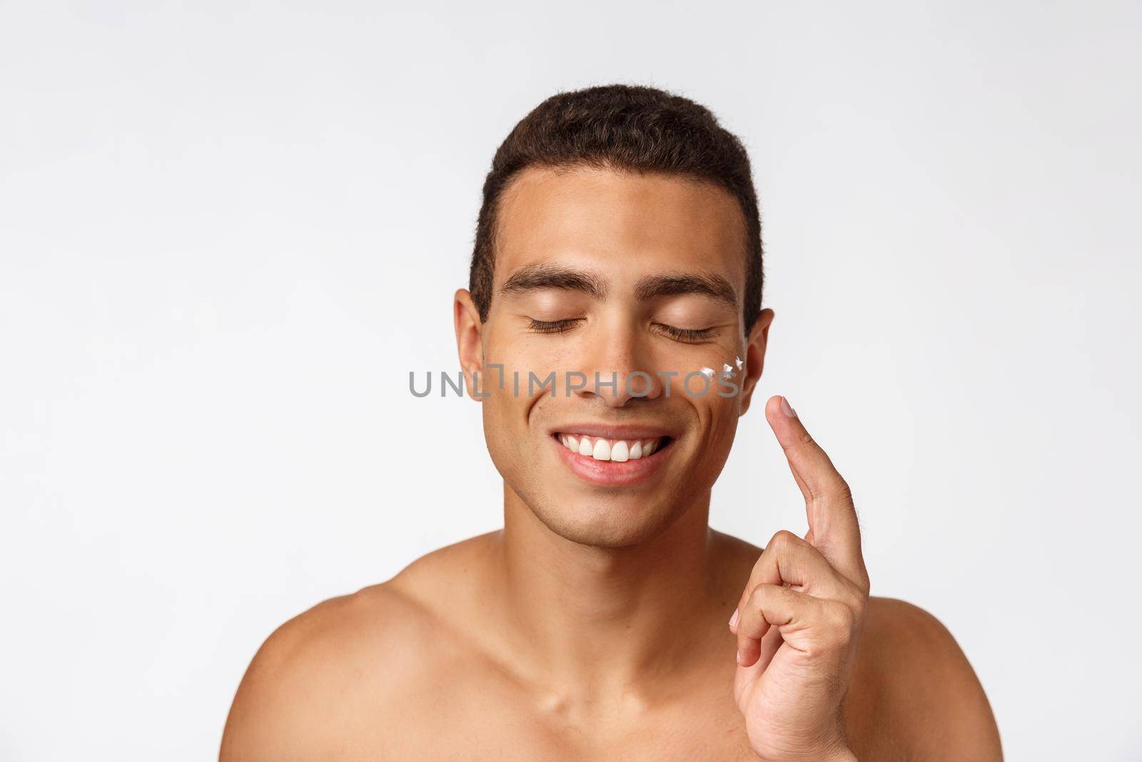 Photo of shirtless african american man smiling and applying face cream isolated over white background.