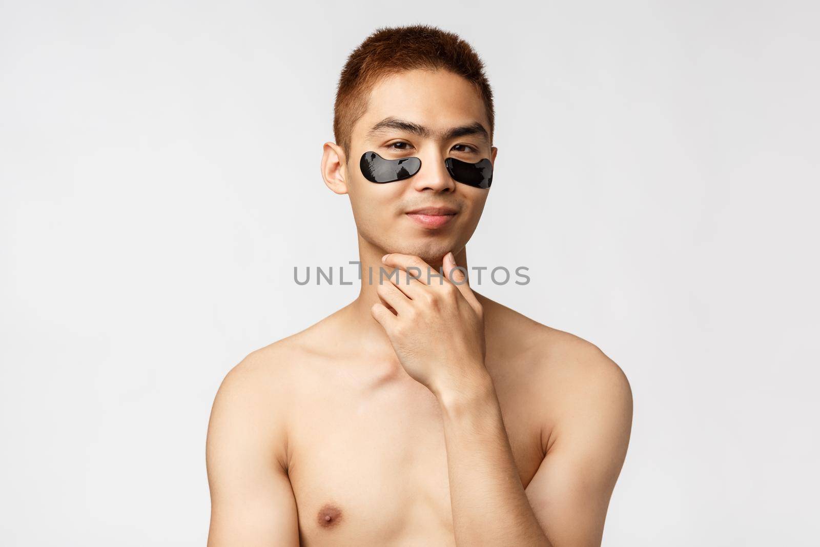 Beauty, people and lifestyle concept. Thoughtful enthusiastic, handsome naked asian man wearing eye-patches and smiling, have an idea, thinking, consider apply skincare mask, white background.