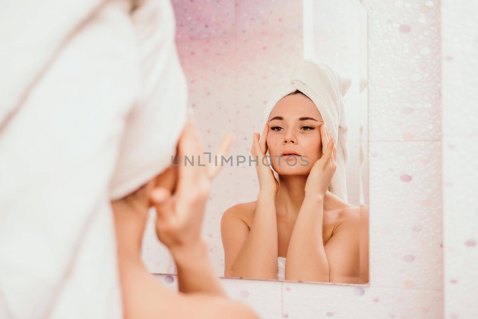 Young beautiful woman using a moisturizing facial mask after taking a bath. Pretty attractive girl in a towel on her head stands in front of a mirror in a home bathroom. Daily hygiene and skin care by Matiunina