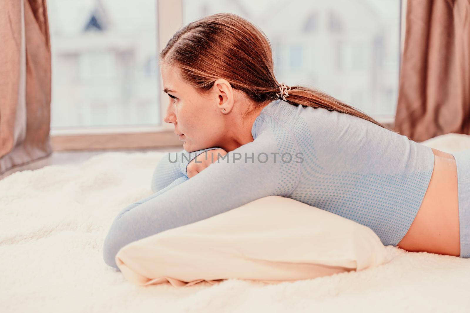 Side view portrait of relaxed woman with long hair lying on carpet at home. She is dressed in a blue tracksuit, holding a phone in her hands