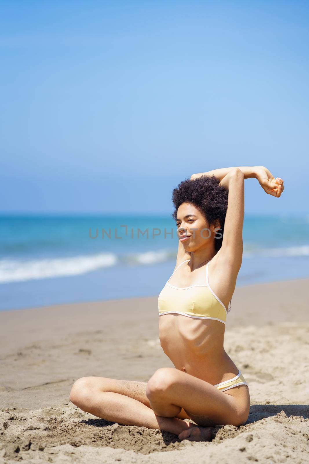 Black woman, wearing a yellow bikini, sitting on the beach relaxing and stretching her body.