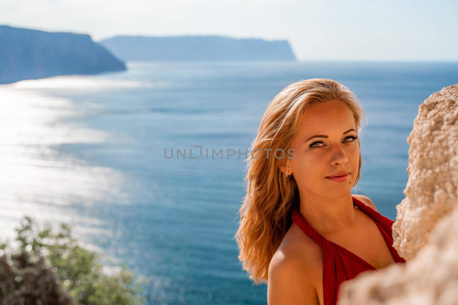 Smiling young woman in a red dress looks at the camera. A beautiful tanned girl enjoys her summer holidays at the sea. Portrait of a stylish carefree woman laughing at the ocean. by Matiunina