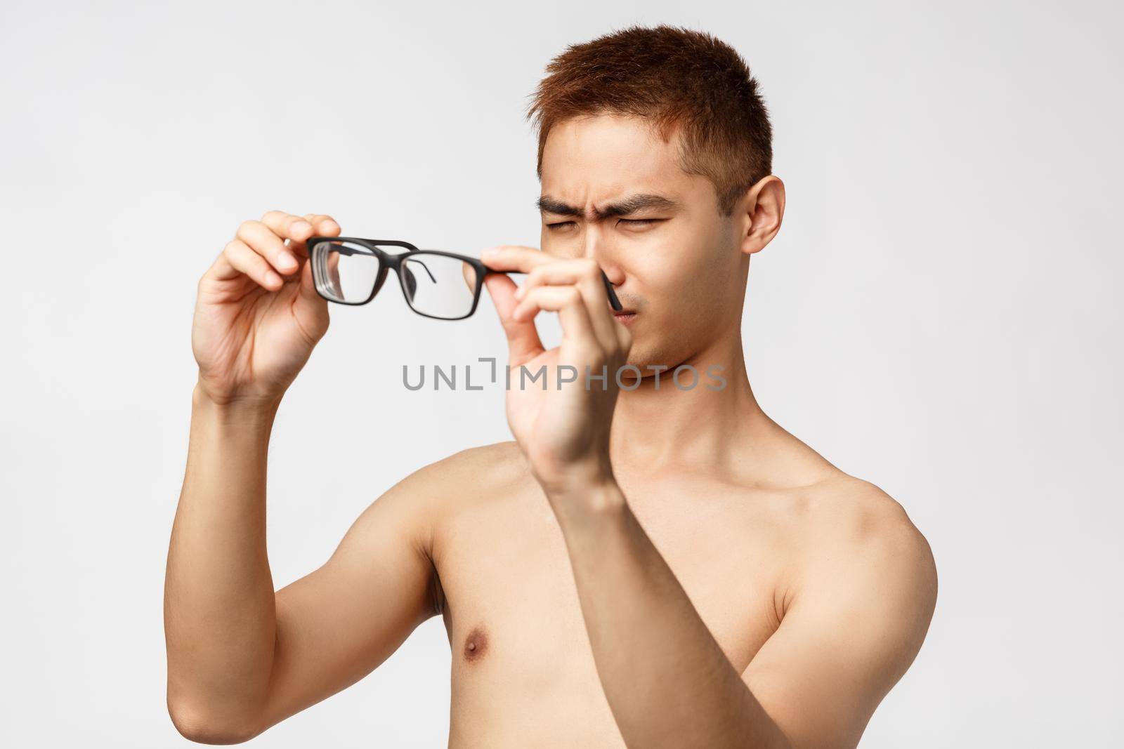 Beauty, people and lifestyle concept. Portrait of young handsome asian man with naked torso, waking up morning cant see anything without glasses, squinting put on eyewear, white background.
