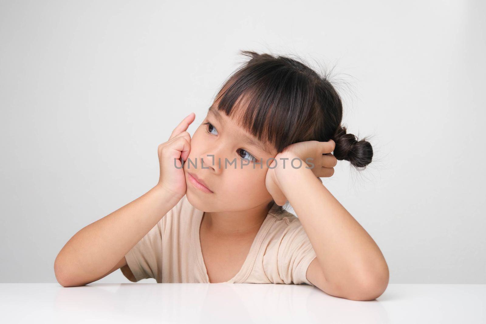Cute dark haired little girl making a thoughtful pose sitting at a table on a white background.