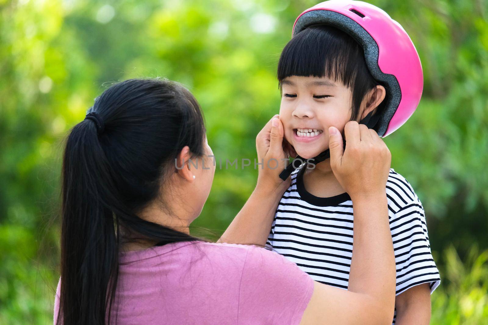 Asian mother helps daughter put on protective pads and safety helmet before practicing roller skating in the park. Exciting outdoor activities for kids. by TEERASAK