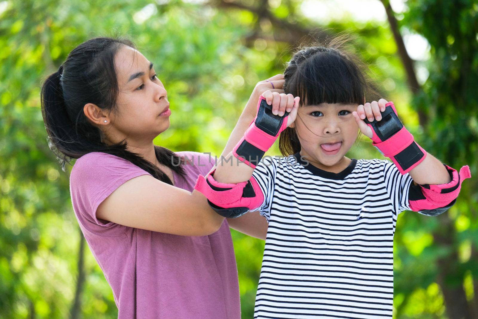 Asian mother helps daughter put on protective pads and safety helmet before practicing roller skating in the park. Exciting outdoor activities for kids. by TEERASAK