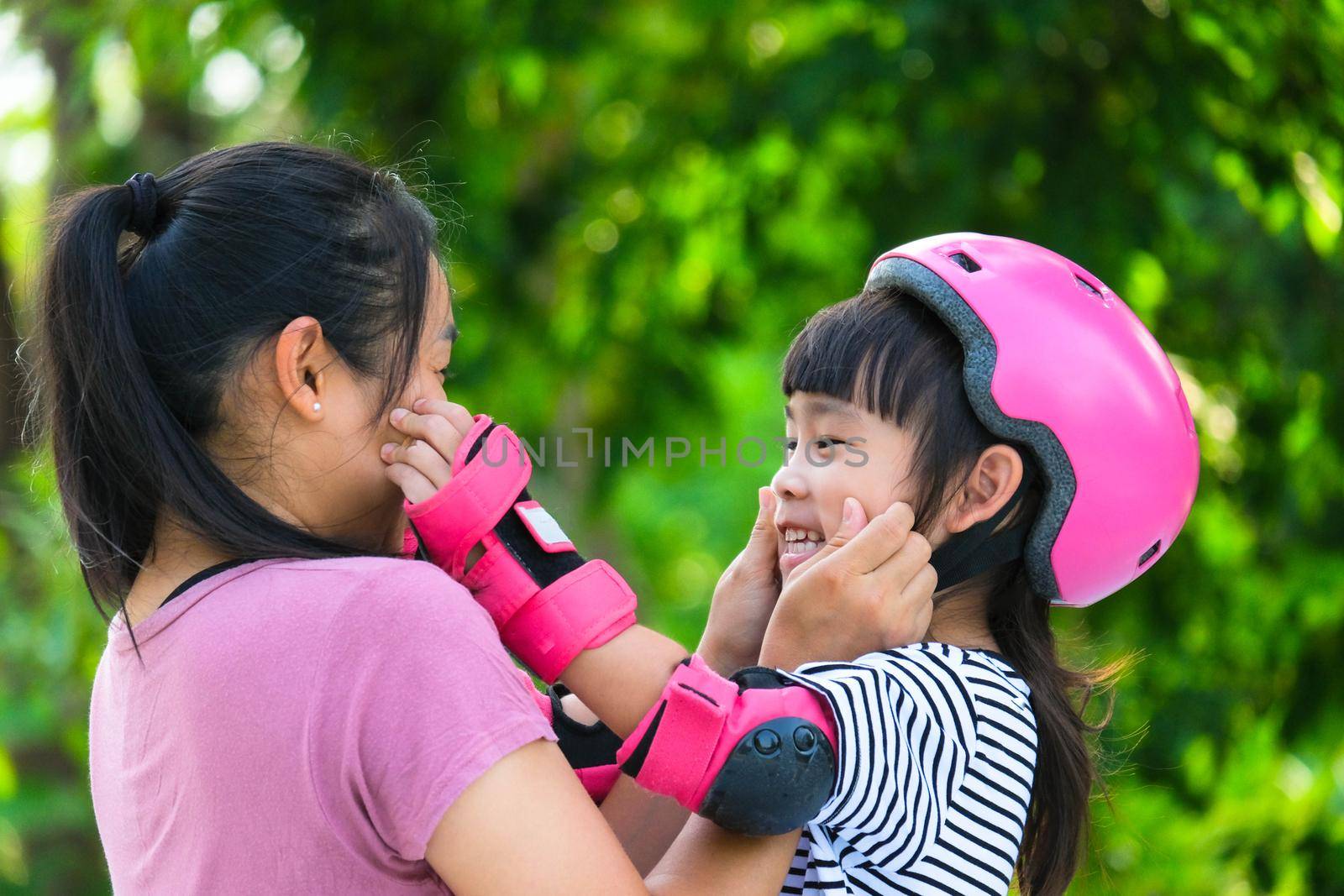 Asian mother helps daughter put on protective pads and safety helmet before practicing roller skating in the park. Exciting outdoor activities for kids.