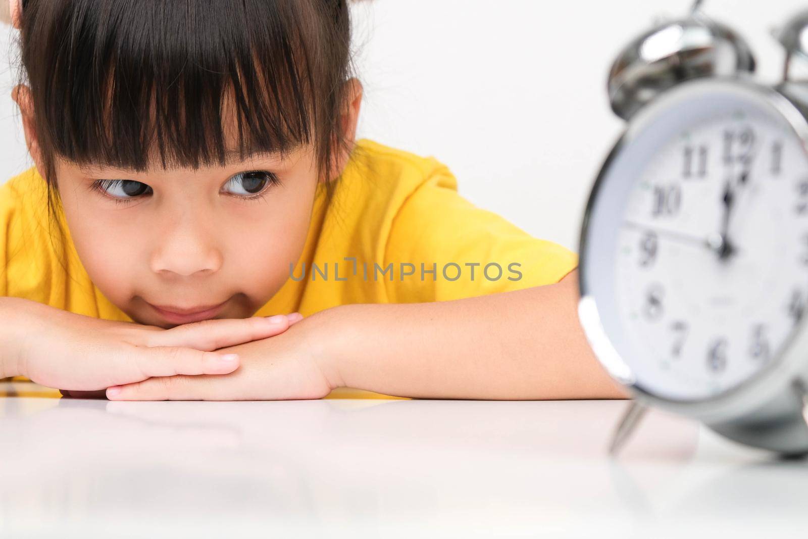 Cute dark haired girl puts her hand under her chin and looks at a vintage alarm clock while sitting at a table on a white background.