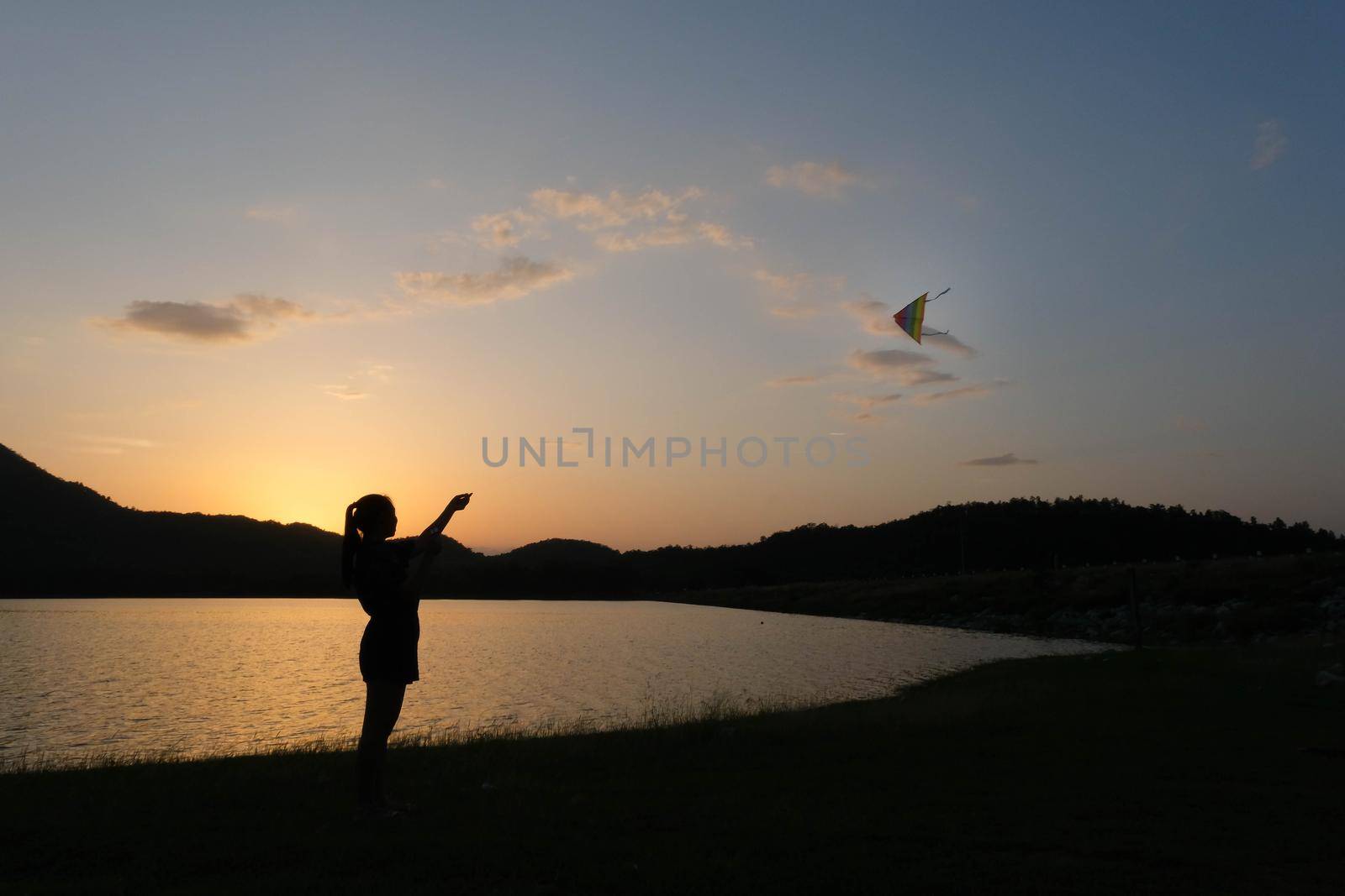 Silhouette of young woman playing a kite by the lake at sunset. by TEERASAK
