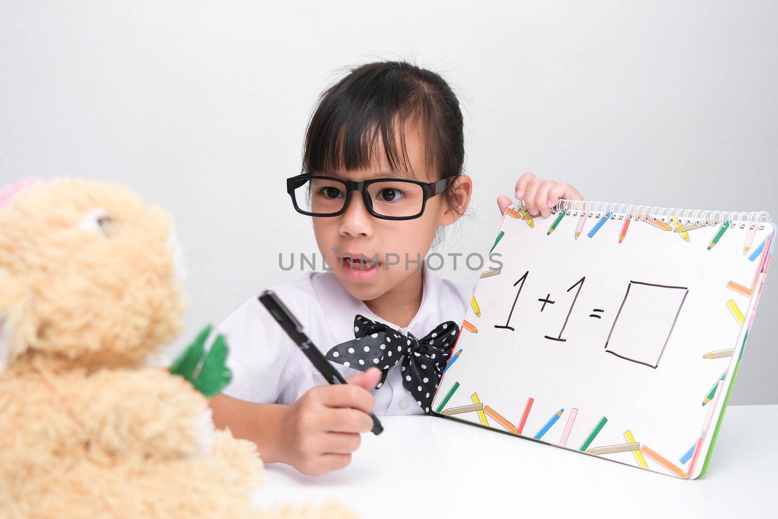 Cute Asian little girl playing teacher role game. A little girl is teaching math to her teddy bear friend. Homeschool children's play and learning. by TEERASAK