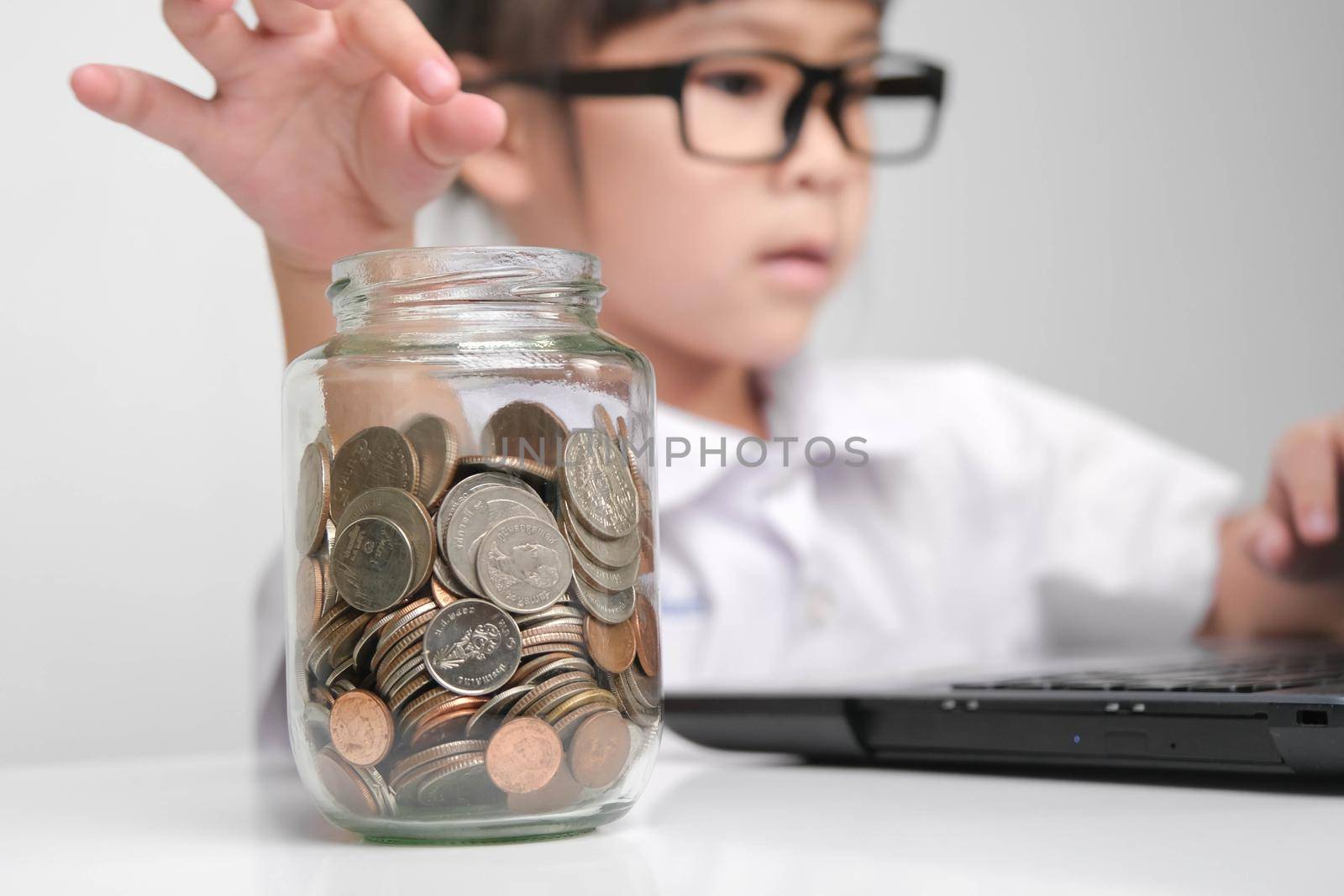 Little businesswoman puts a coin in a glass jar on a table while working with laptop in office. Children and business concept by TEERASAK