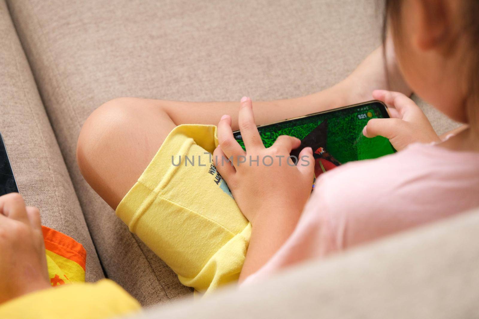 Two little girls addicted to mobile phones. Asian sisters are playing games or watching cartoons with their smartphones on the sofa at home. by TEERASAK