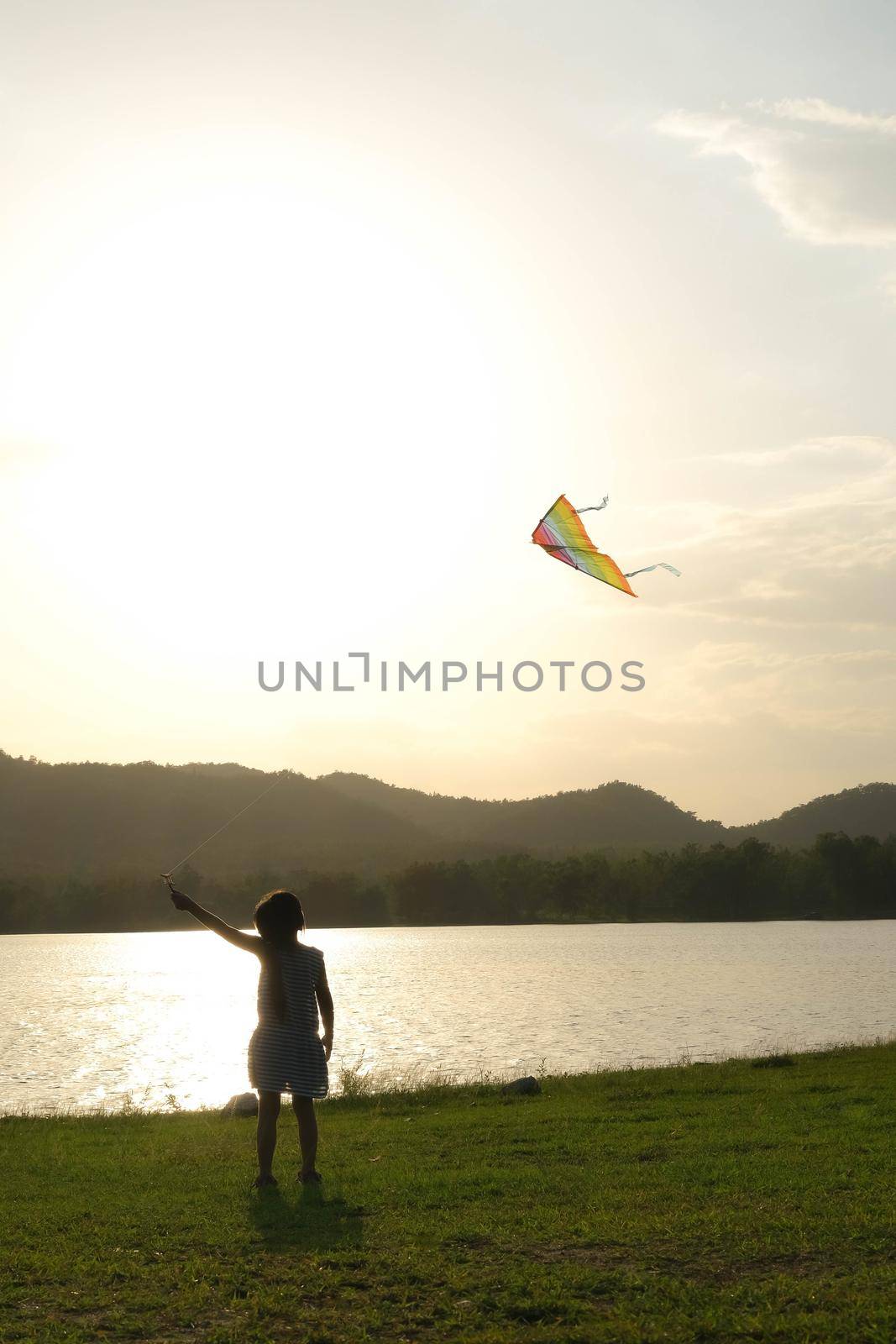 Child playing with a kite while running on a meadow by the lake at sunset. Healthy summer activity for children. Funny time with family. by TEERASAK