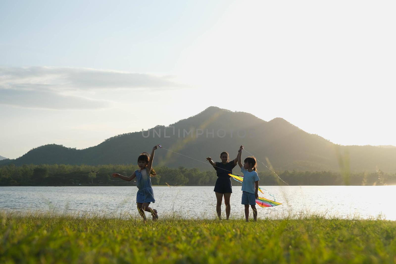 Children are flying kites while running on a meadow by the lake at sunset with their mother. Healthy summer activity for children. Funny time with family. by TEERASAK