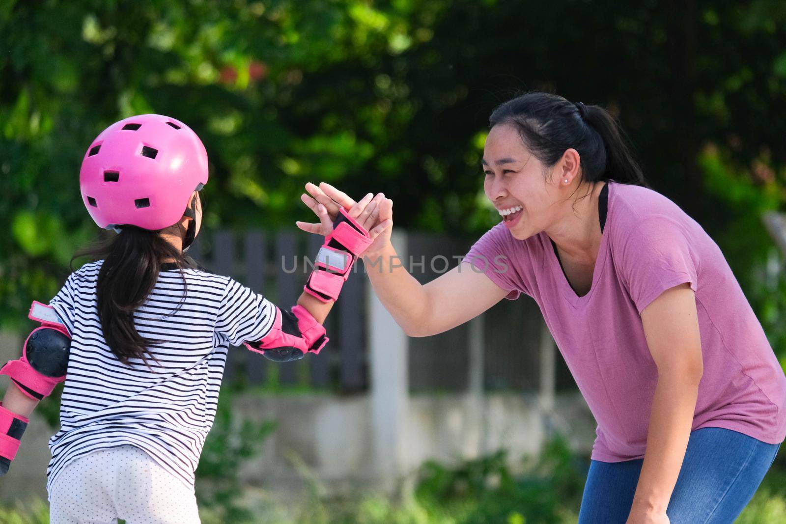 Asian mother helps daughter practice roller skating in the park. Exciting outdoor activities for kids. by TEERASAK