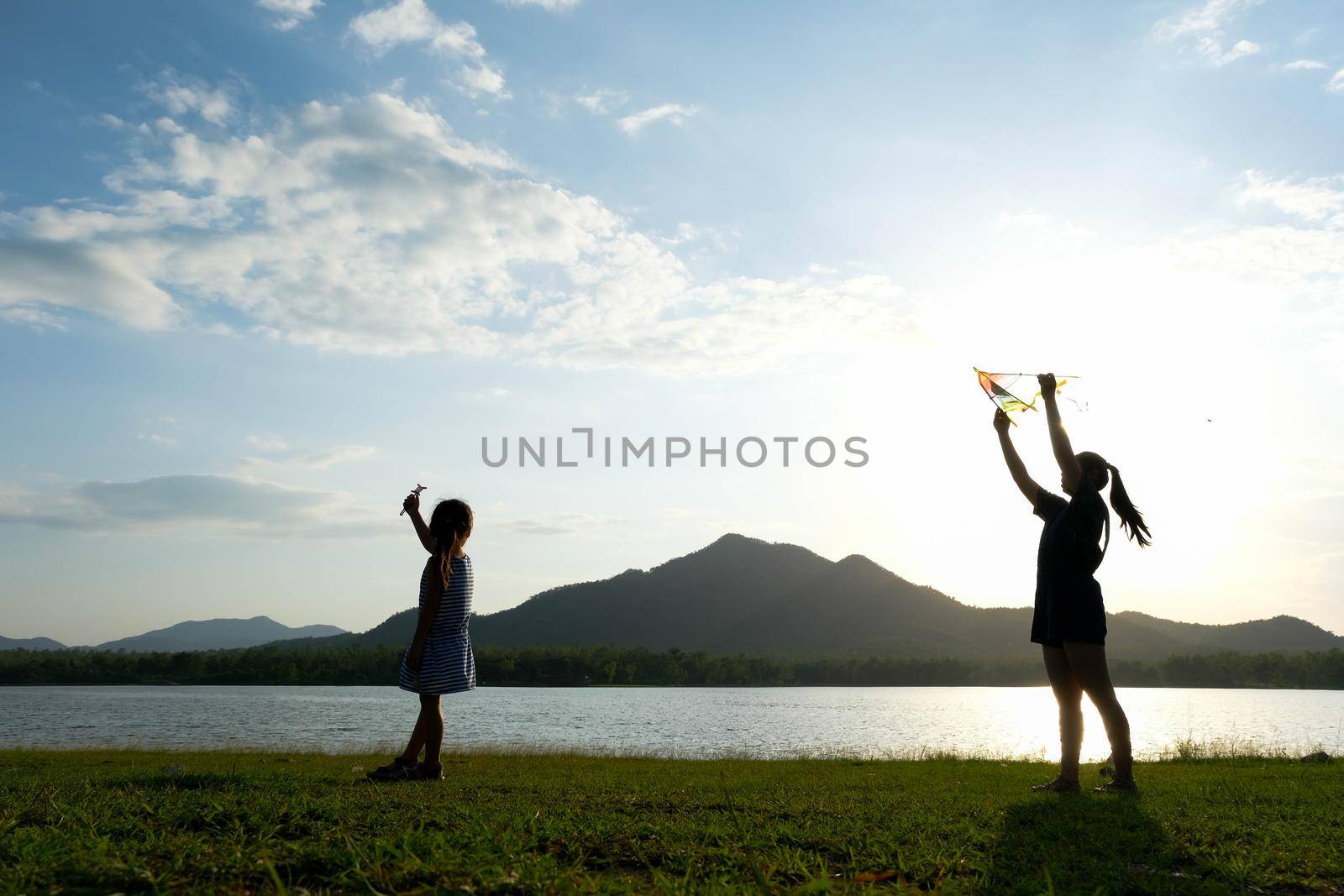 Children are flying kites while running on a meadow by the lake at sunset with their mother. Healthy summer activity for children. Funny time with family. by TEERASAK