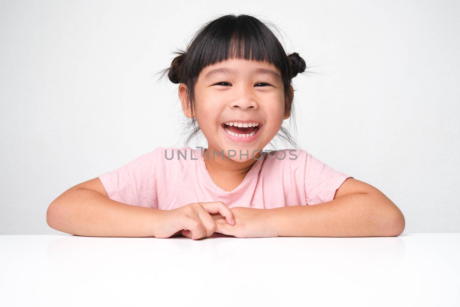 Cute dark haired girl smiling happily sitting at a table on a pink background and looking at the camera. Advertising childrens products