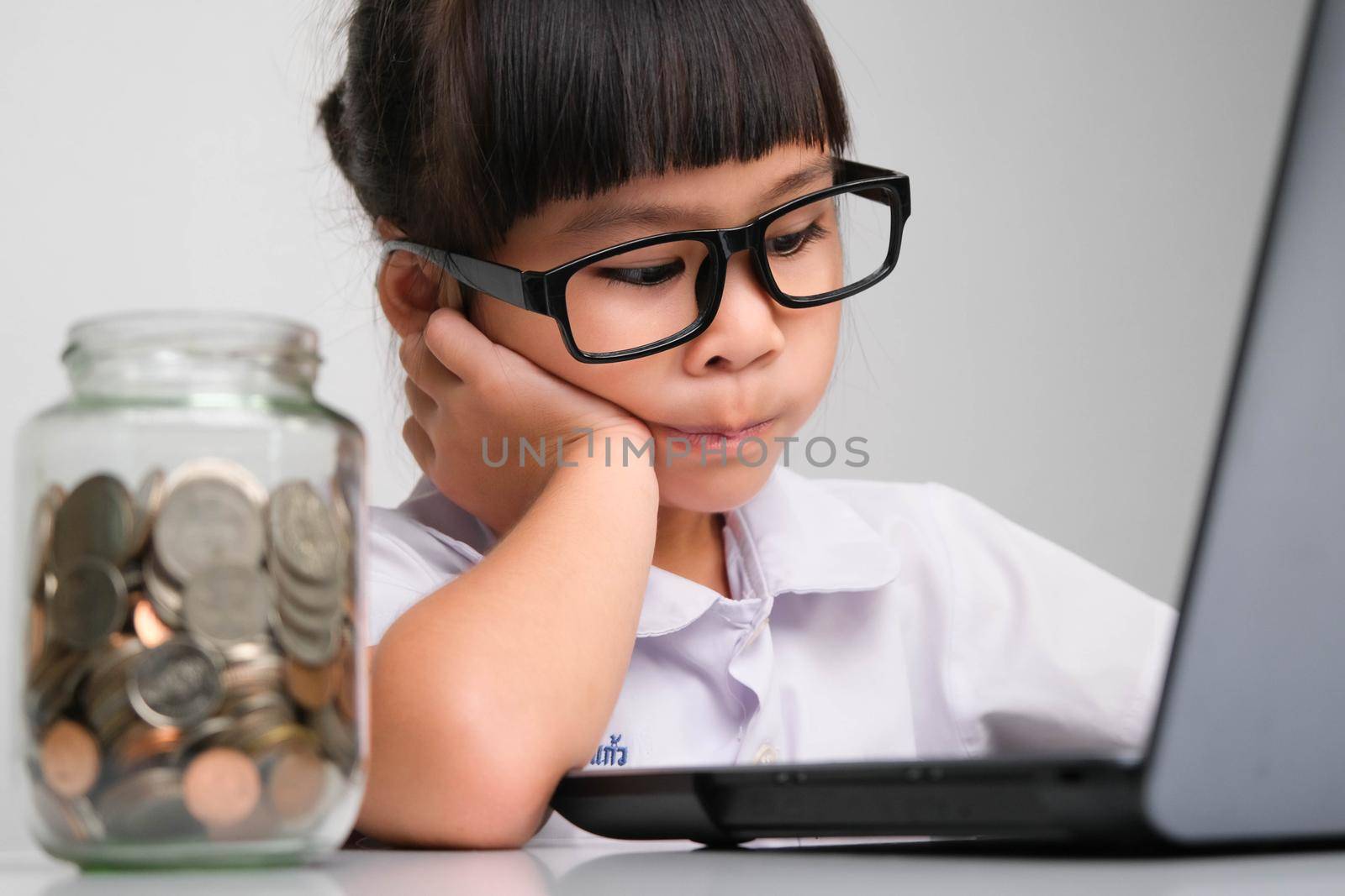 Little businesswoman with laptop working in office with coins in a glass jar on the table. Children and business concept by TEERASAK