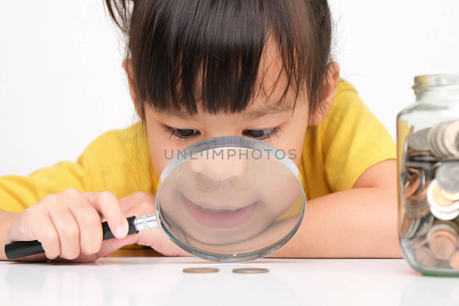 Cute Asian little girl holding a magnifying glass looking at coins on the table and a clear jar full of coins near her. Children learning about saving for future concept by TEERASAK