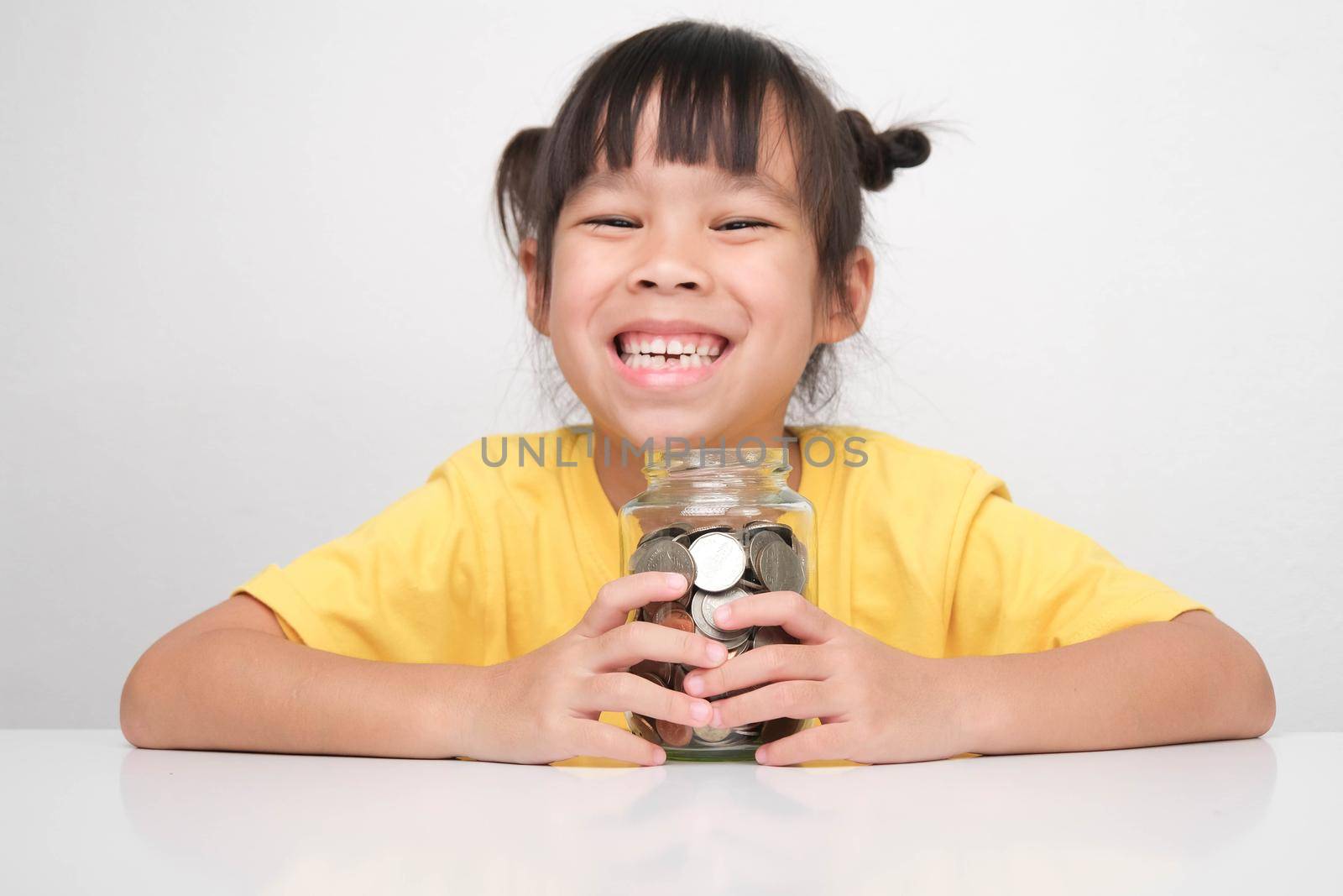 Cute Asian little girl smiling happily holding money coins in clear jar sitting at a table on white background and looking at the camera. Children learning about saving for future concept by TEERASAK
