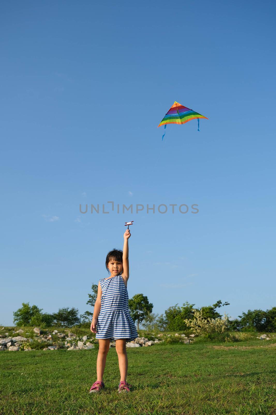 Child playing with a kite while running on a meadow by the lake at sunset. Healthy summer activity for children. Funny time with family. by TEERASAK