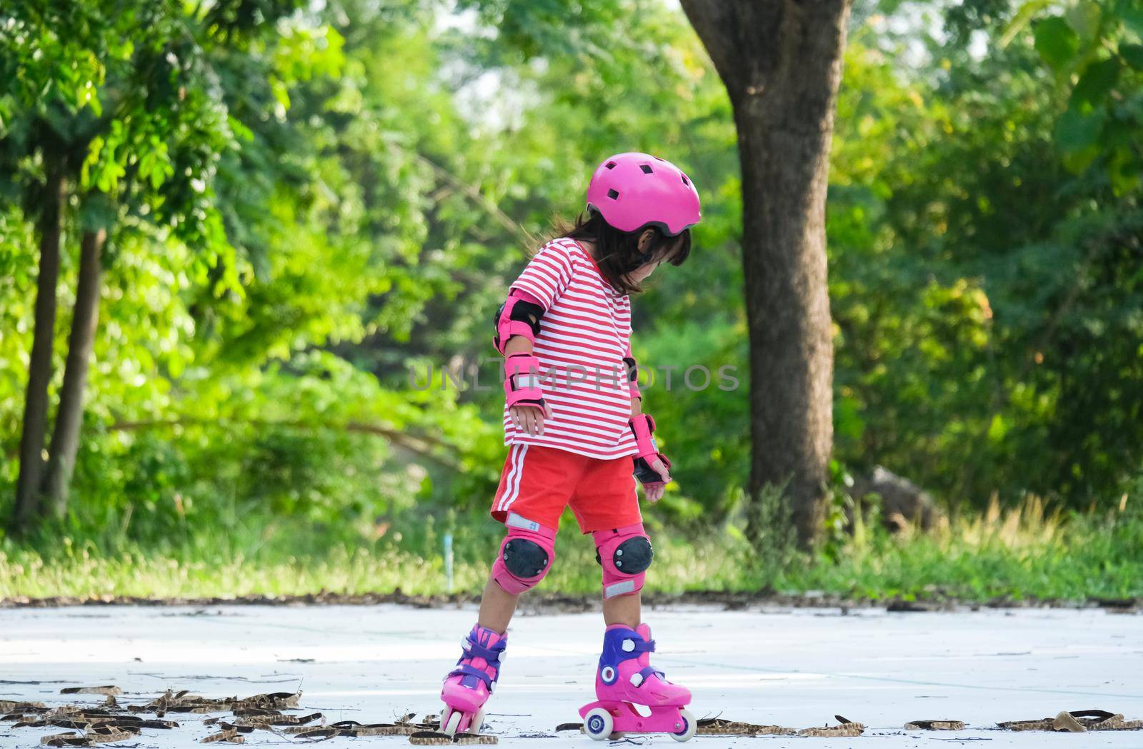 Cute Asian little girl in protective pads and safety helmet practicing roller skating in the park. Exciting outdoor activities for kids. by TEERASAK