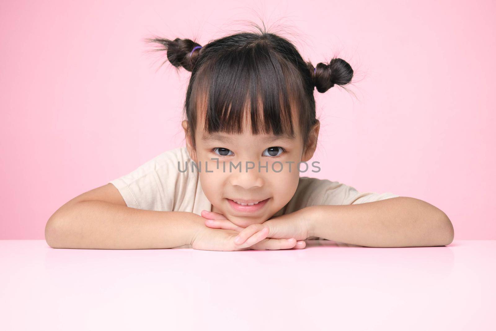 Cute dark hair little girl smiles happily and puts her hand under her chin while sitting at a table on a pink background and looking at the camera. Advertising childrens products by TEERASAK
