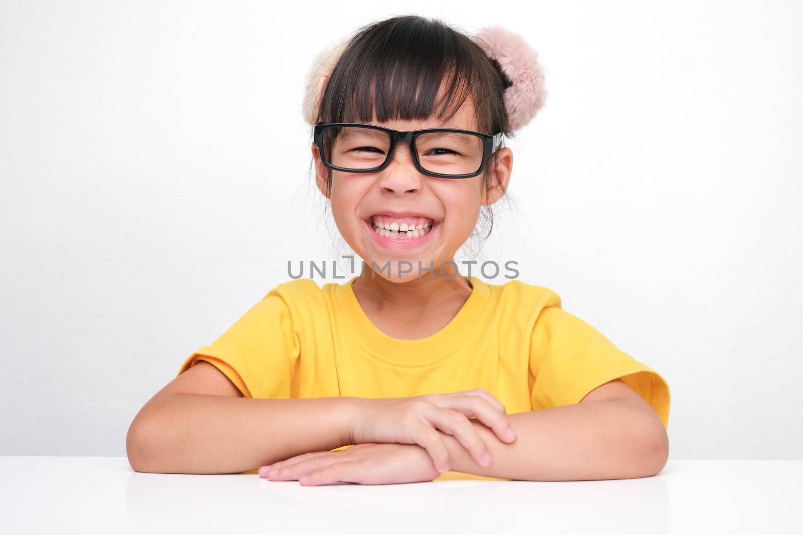 Cute dark haired girl smiling happily sitting at a table on white background and looking at the camera. Advertising childrens products