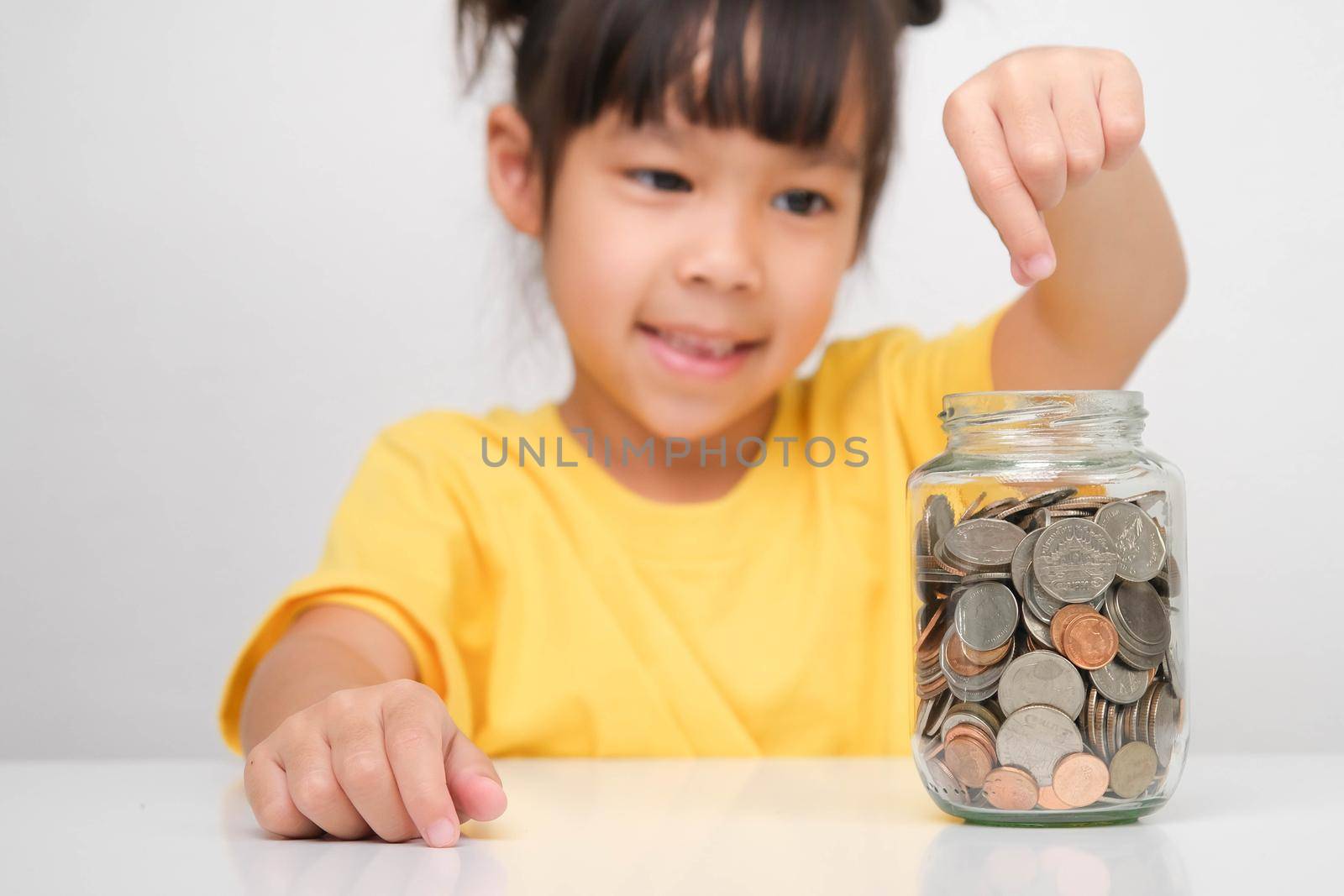 Happy smiling cute Asian girl holding a coin and putting it into a clear jar sitting at a table on a white background. Children learning about saving for future concept by TEERASAK