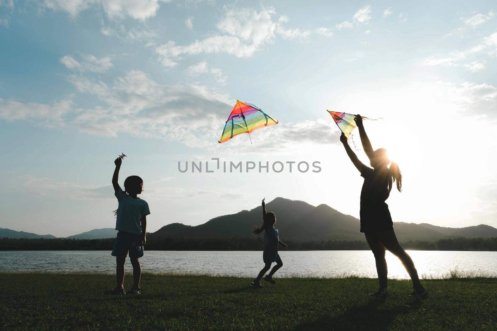 Children are flying kites while running on a meadow by the lake at sunset with their mother. Healthy summer activity for children. Funny time with family. by TEERASAK