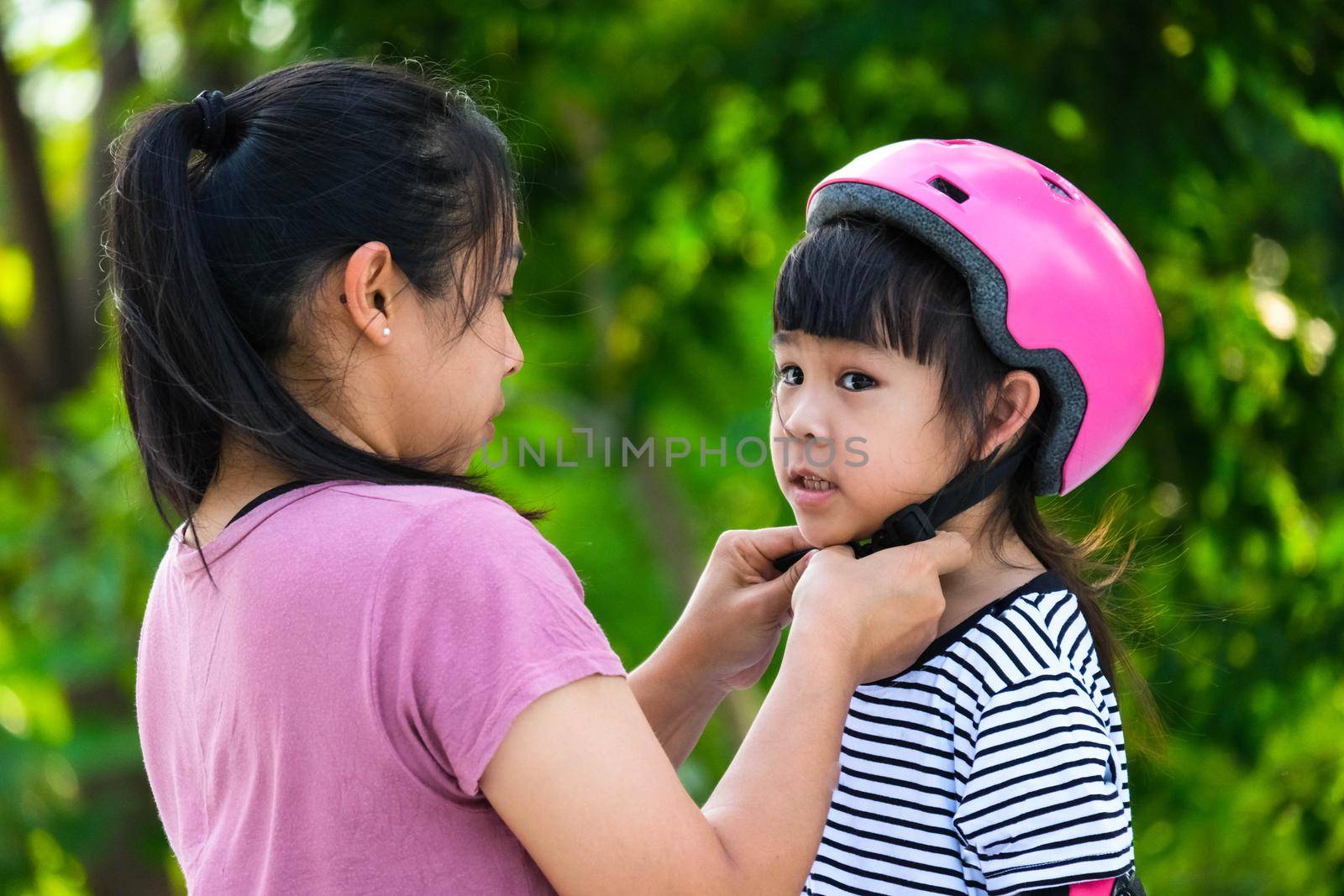 Asian mother helps daughter put on protective pads and safety helmet before practicing roller skating in the park. Exciting outdoor activities for kids.