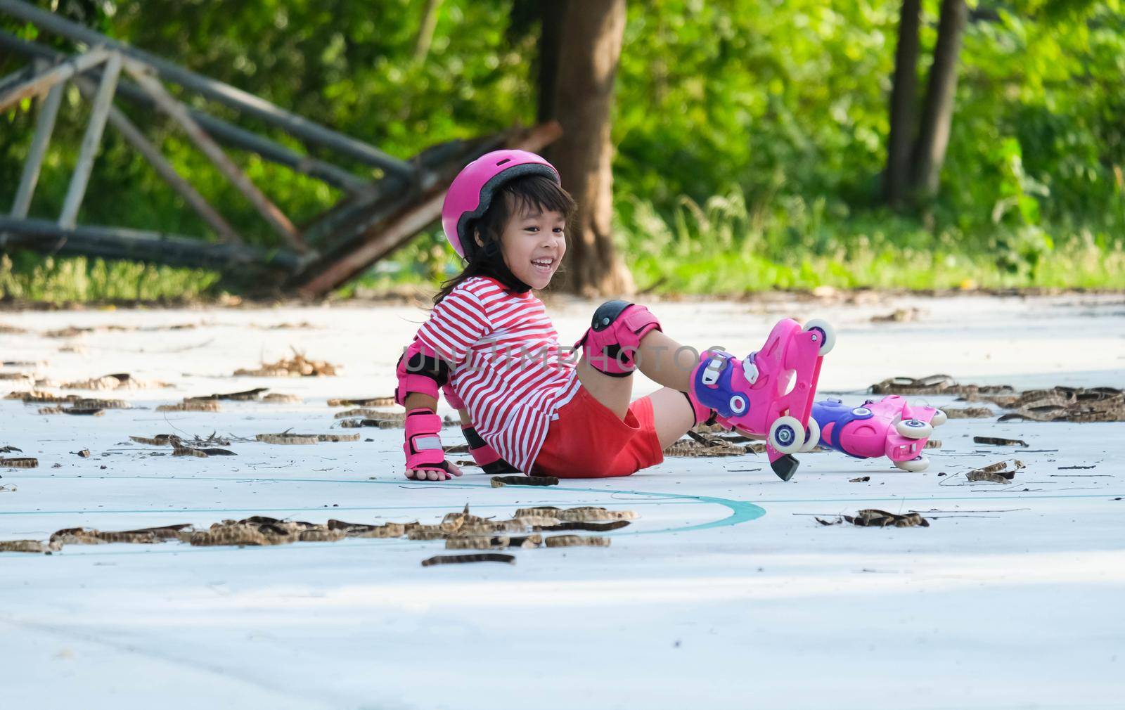 Cute Asian little girl in protective pads and safety helmet practicing roller skating in the park. Exciting outdoor activities for kids. A preschooler wearing roller skates falls down. by TEERASAK
