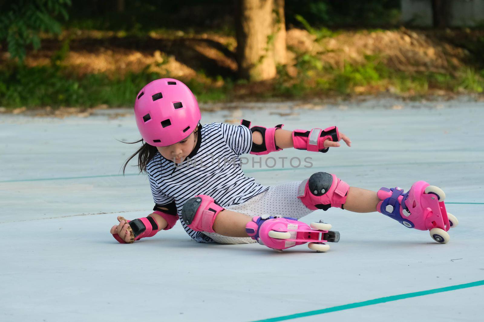 Cute Asian little girl in protective pads and safety helmet practicing roller skating in the park. Exciting outdoor activities for kids. A preschooler wearing roller skates falls down. by TEERASAK