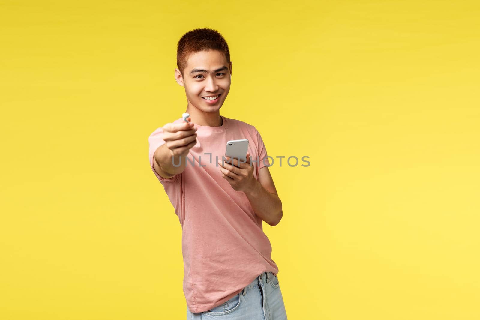 Technology, communication and lifestyle concept. Portrait of young stylish asian guy giving wireless headphone to you, sharing earphones to listen music together, hold mobile phone.