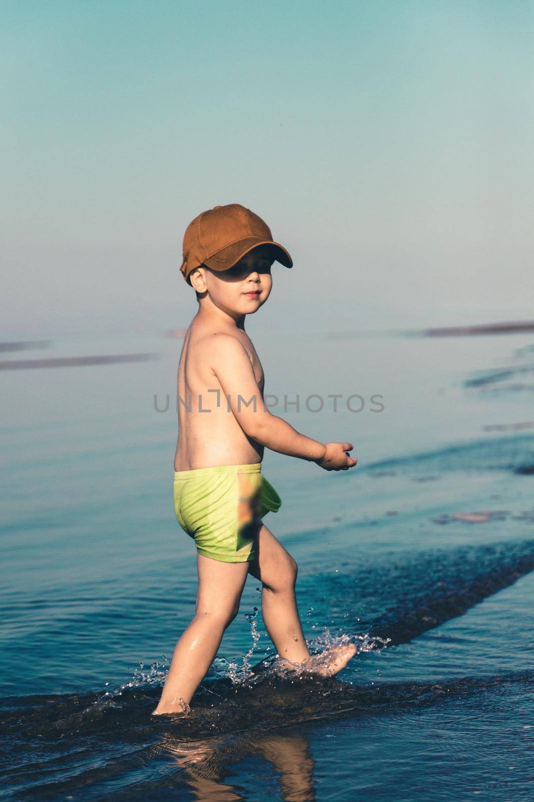 A boy in green shorts and a brown baseball cap walks on the sea near the shore. Copy space. Toned minimalism vertical photo.