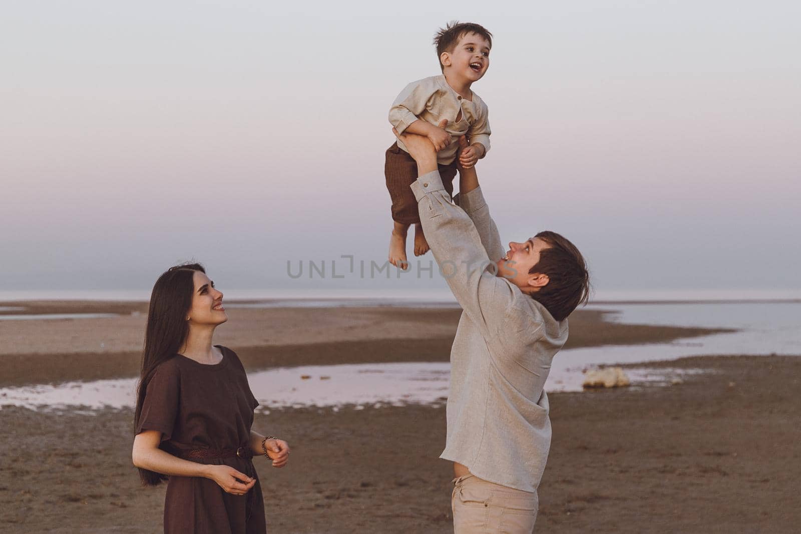 Young family playing with their child on the evening beach by the sea. Father fun throws up son in the air.