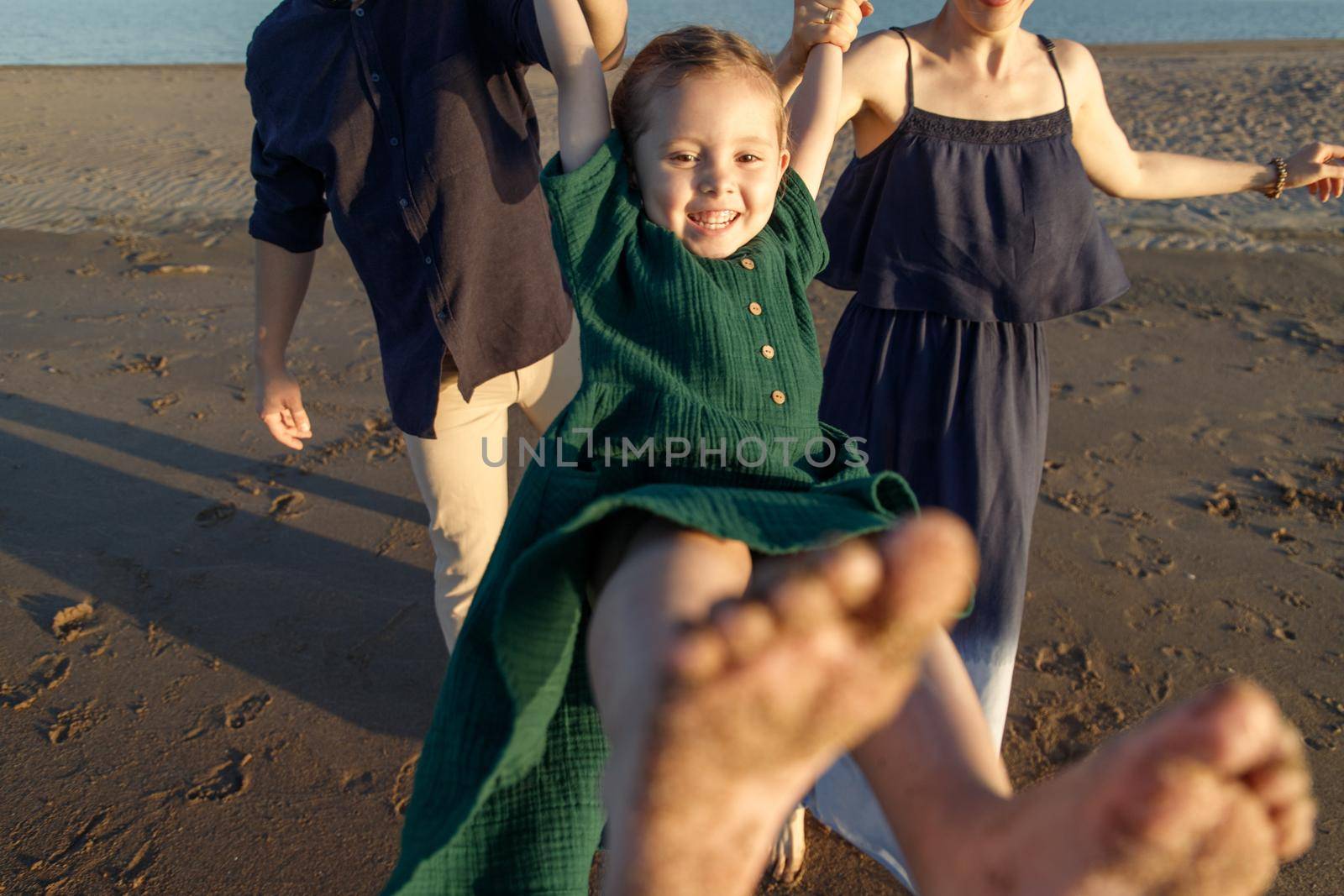 Fun portrait of a happy girl in a green dress swinging in the arms of her parents outdoor.