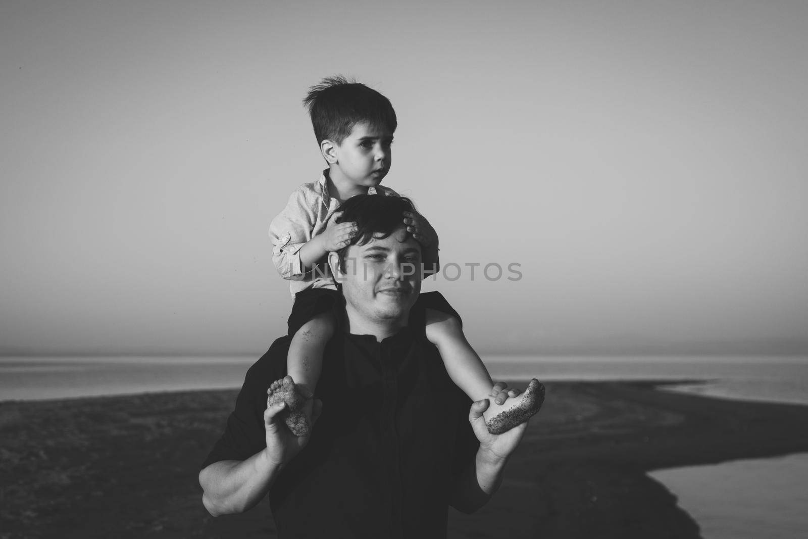 Black and white portrait of a cute boy 3 years old on his father's neck walking on the beach in sunset lighting.