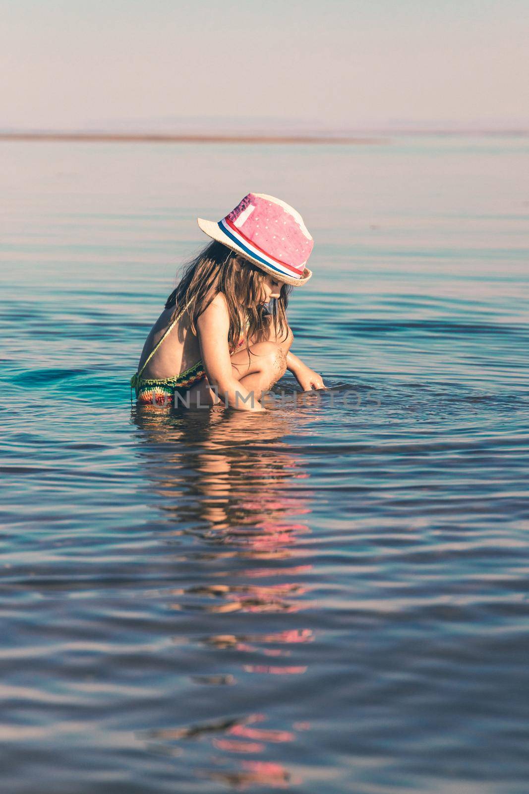 Cute little girl in a knitted swimsuit and a pink hat is playing with water while sitting in the sea near the shore. Copy space. Toned minimalism vertical
