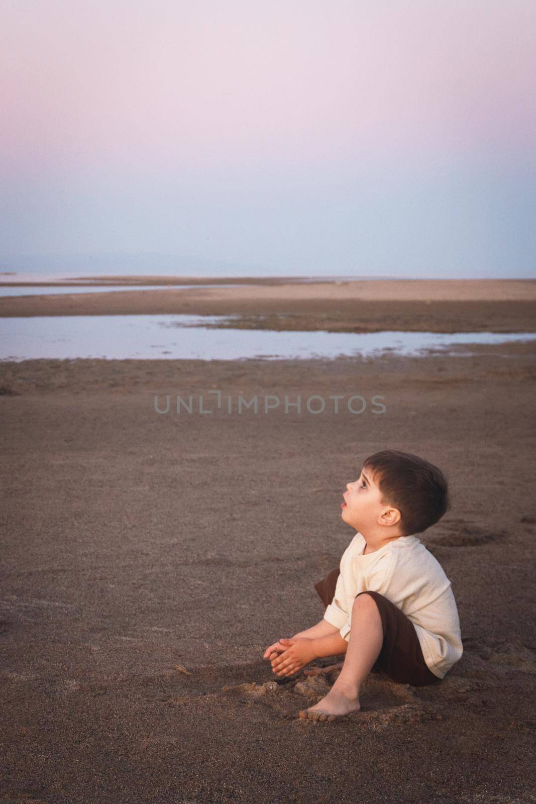 Little cute boy 3 years old plays with sand on the beach in the evening, he is look up. Vertical, copy space.