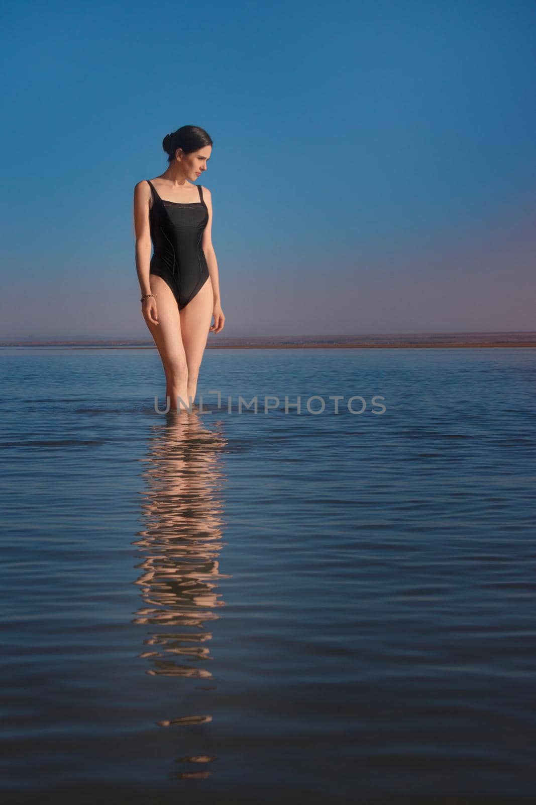 Young beautiful slim woman wearing a black swimsuit stands in the sea and watching sunset. Vertical by Rom4ek