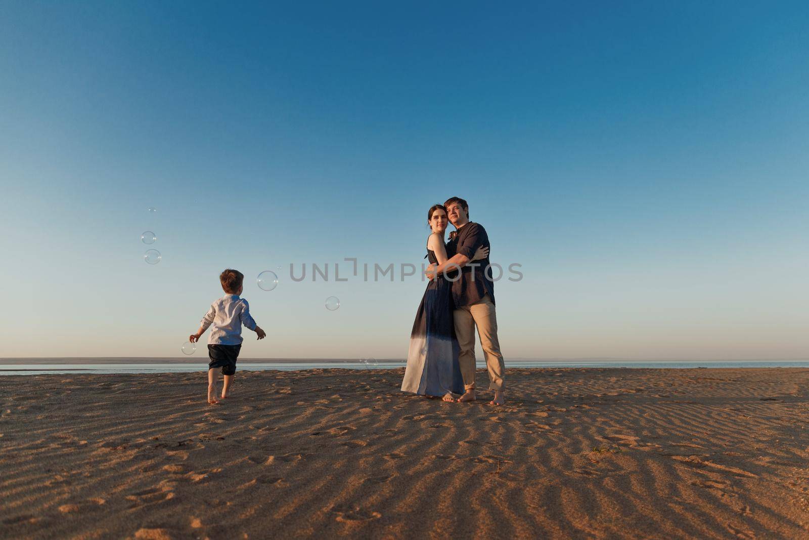 Young couple with their little son are walking along the beach in the evening natural light, soap bubbles fly around by Rom4ek