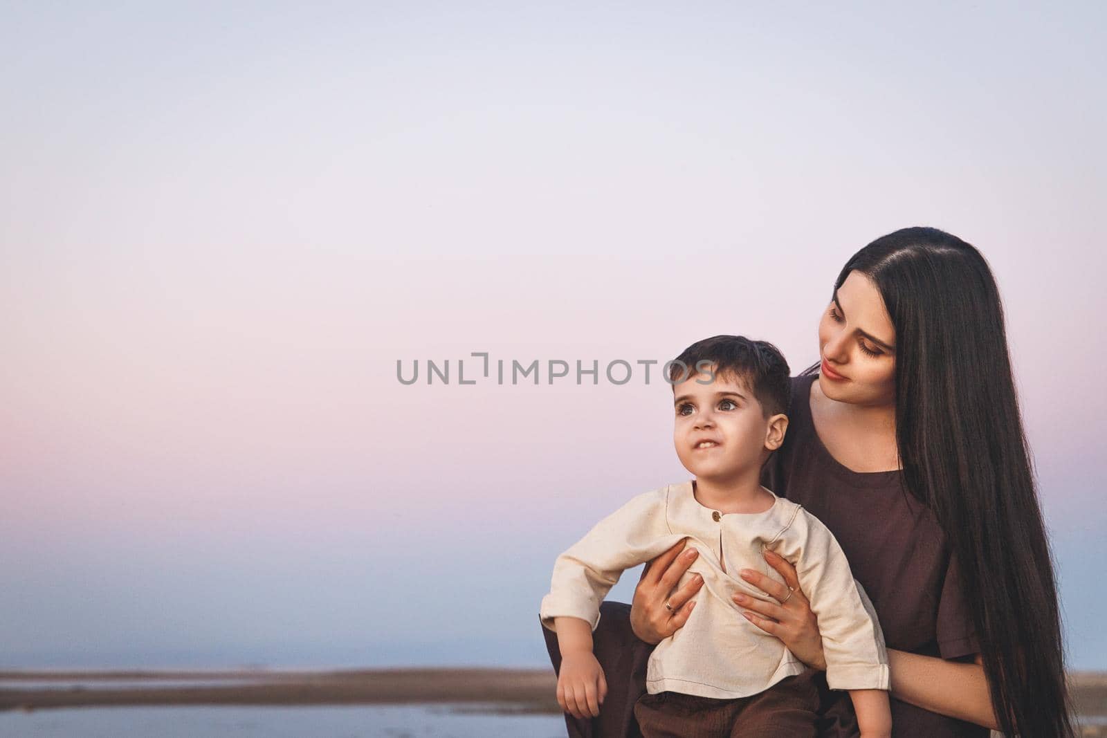 Happy young beautiful mother with her son three years old admiring the sunset on the beach. Family look linen clothes by Rom4ek