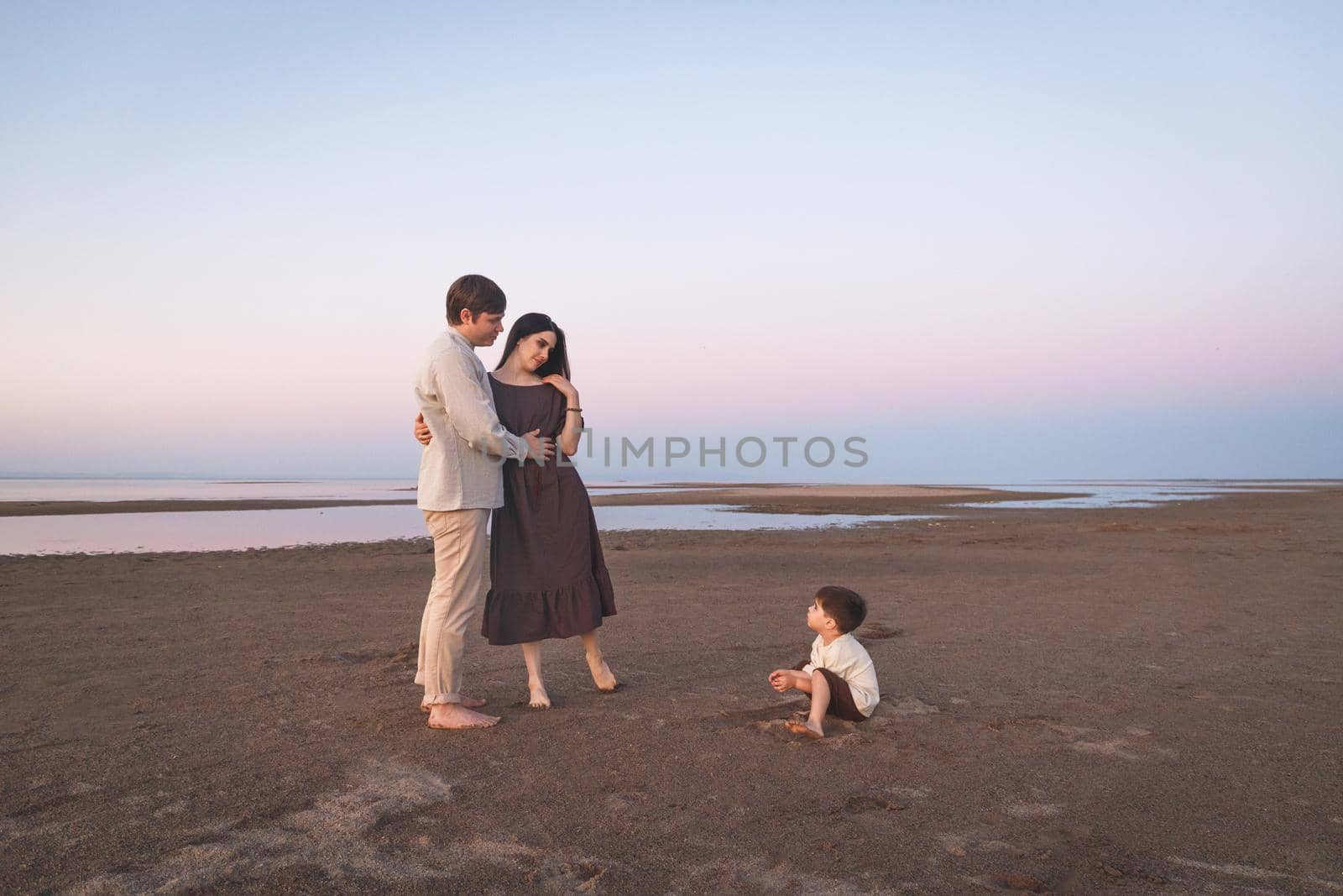 A young family in linen clothes with a 3 year old son is walking along the wild evening beach. Copy space.