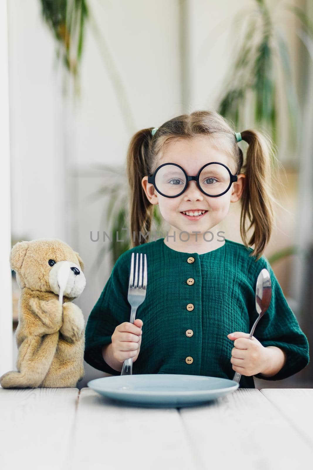 Little beautiful girl in green muslin dress waiting breakfast with bear toy. Little cute girl at white dining table in kitchen. Healthy nutrition for young kids.