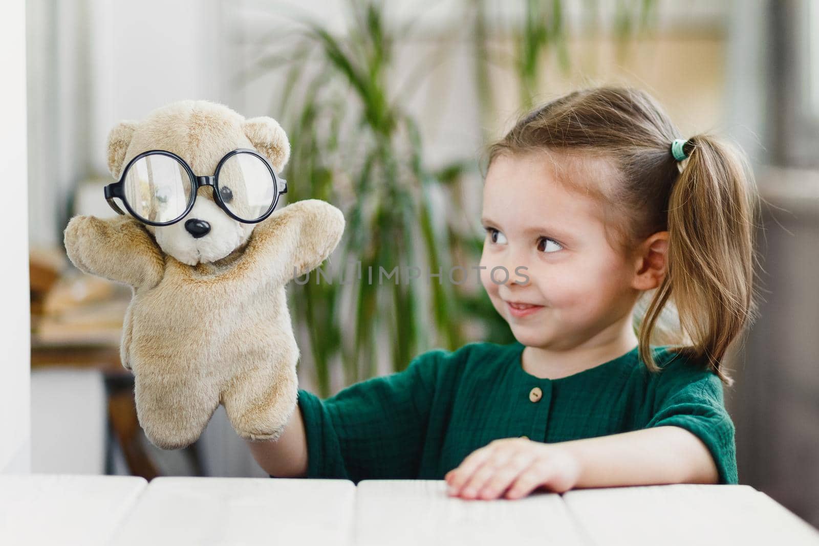 Cute little girl is playing with a teddy bear in round shaped glasses and smiling while stay at home.