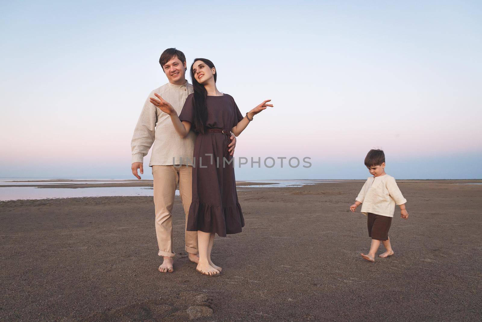 Young family with a three year old son walks barefoot along the wild evening beach. Family look linen clothes.