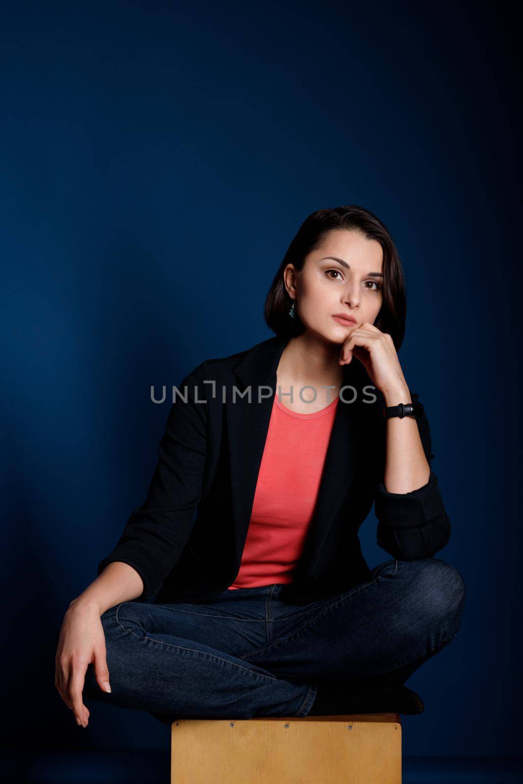 Portrait of young brunette woman wearing a coral tank top, black blazer and blue jeans on dark blue background.