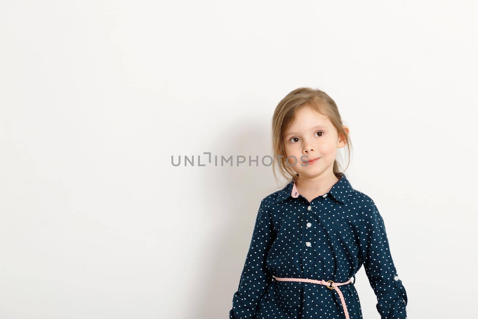 Little cute caucasian girl 4-6 years old wearing a blue dress with polka dots stands against the background of a white wall.