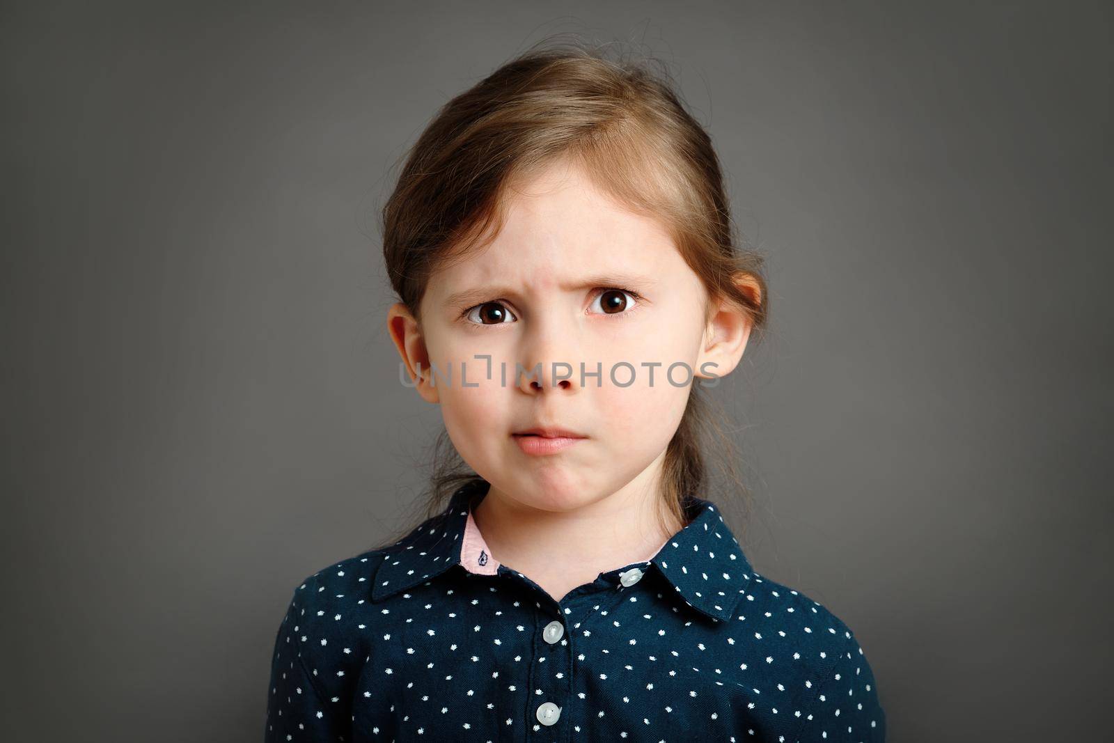 Perplexed little caucasian kid girl 4 -6 years old in a blue dress with polka dots on grey background studio portrait by Rom4ek